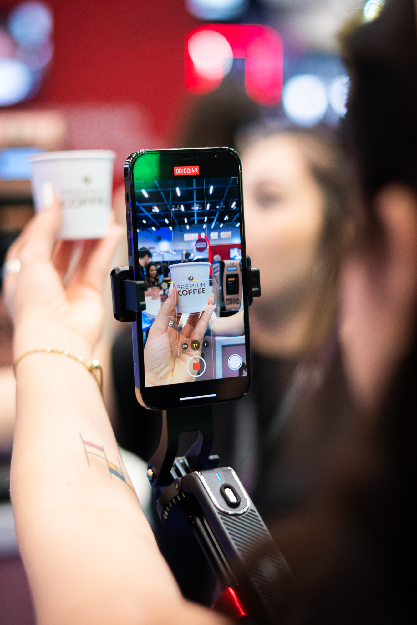 Imagem de Sophia Ferreira, UGC Creator, capturando um momento de produção de conteúdo no Evento Influent Summit. Na foto, um smartphone montado em um estabilizador grava um vídeo de um copo de café premium. A mão de Sophia Ferreira, modelo e empreendedora, segura o copo, enquanto a tela do celular exibe o enquadramento do vídeo. O ambiente do evento é dinâmico, com um fundo desfocado de pessoas interagindo, destacando a expertise da comunicadora e influencer na criação de conteúdos visuais.