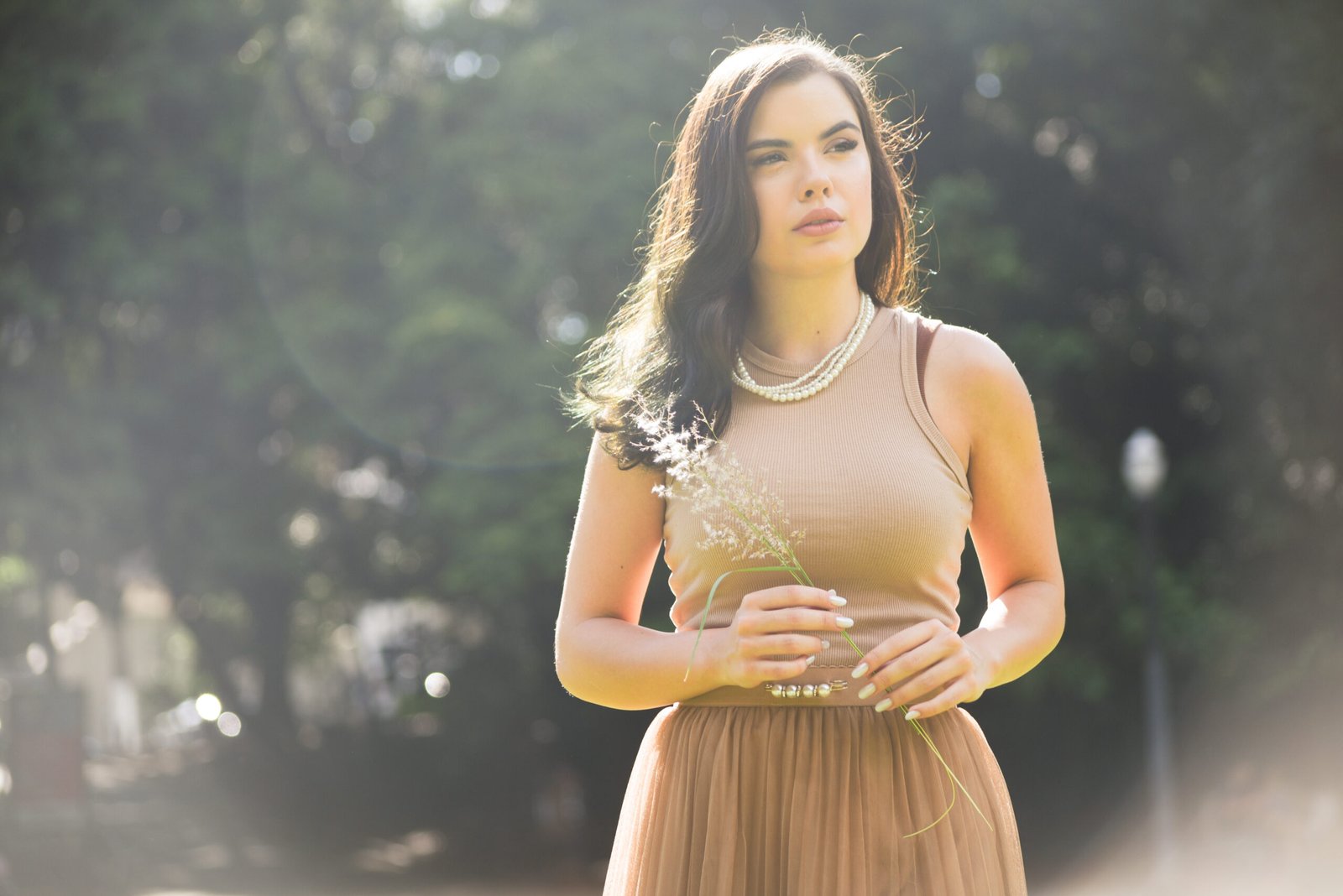 Sophia Ferreira, UGC Creator e criadora de conteúdo digital, posando ao ar livre com um vestido elegante em tons terrosos, segurando uma flor, com luz natural e efeito de flare ao fundo.