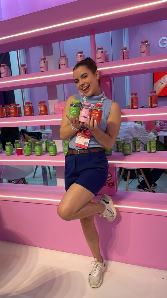 Sophia Ferreira sorridente, segurando potes de gomas vitamínicas Gummy Hair em um stand vibrante, representando a experiência de um criador de conteúdo digital com um produto do segmento de bem-estar.