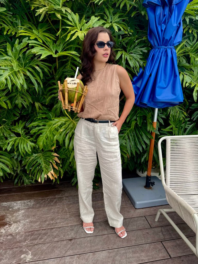 Sophia Ferreira vestindo blusa marrom e calça bege da Amoréia, segurando um coco personalizado em frente a um cenário tropical.