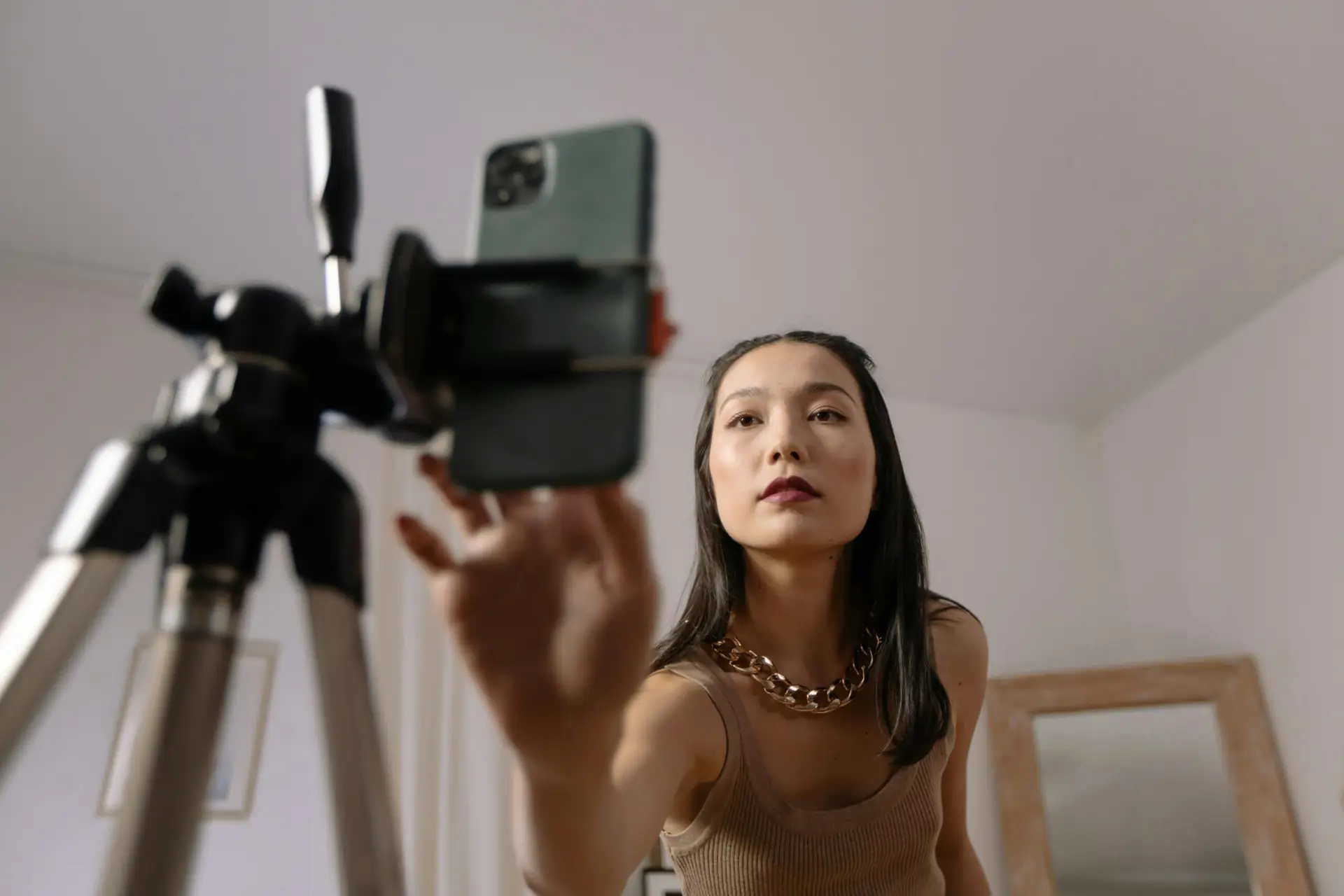 Mulher jovem e profissional ajustando um smartphone em um tripé para gravação de conteúdo digital. O cenário moderno e sofisticado sugere um ambiente de criação de User Generated Content (UGC), tendência crescente no marketing digital e de luxo.