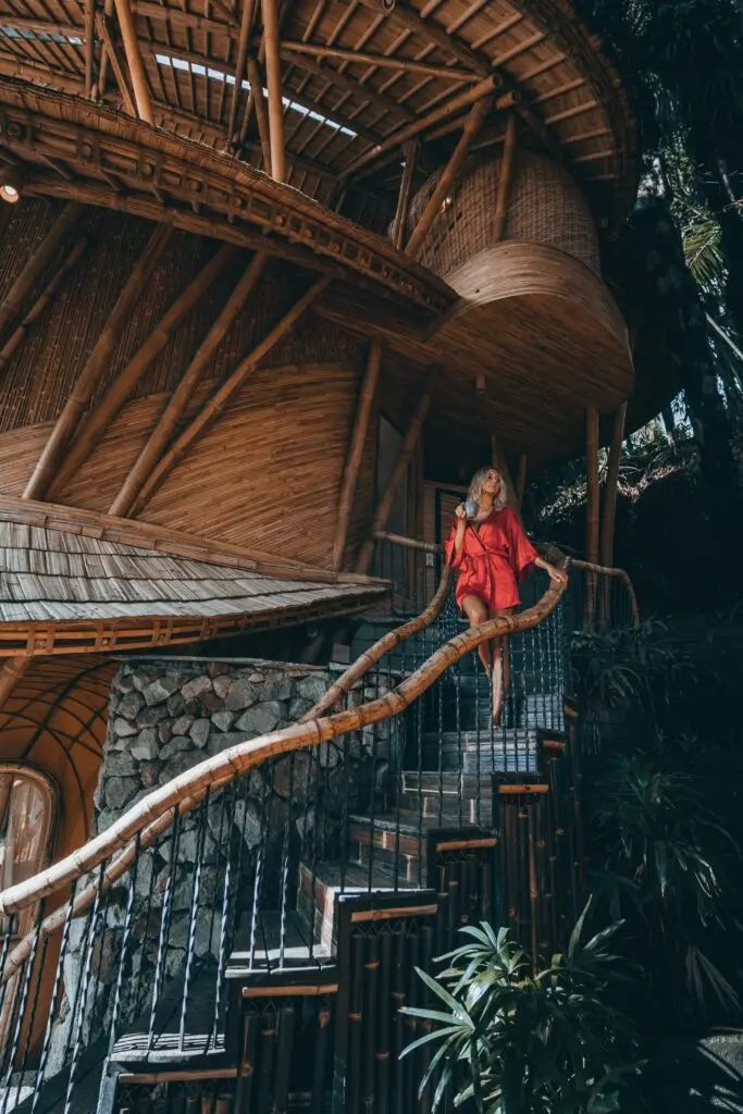 Mulher caminhando por uma construção ecológica feita de bambu, vestindo um vestido vermelho vibrante, representando luxo sustentável e estilo de vida exclusivo.