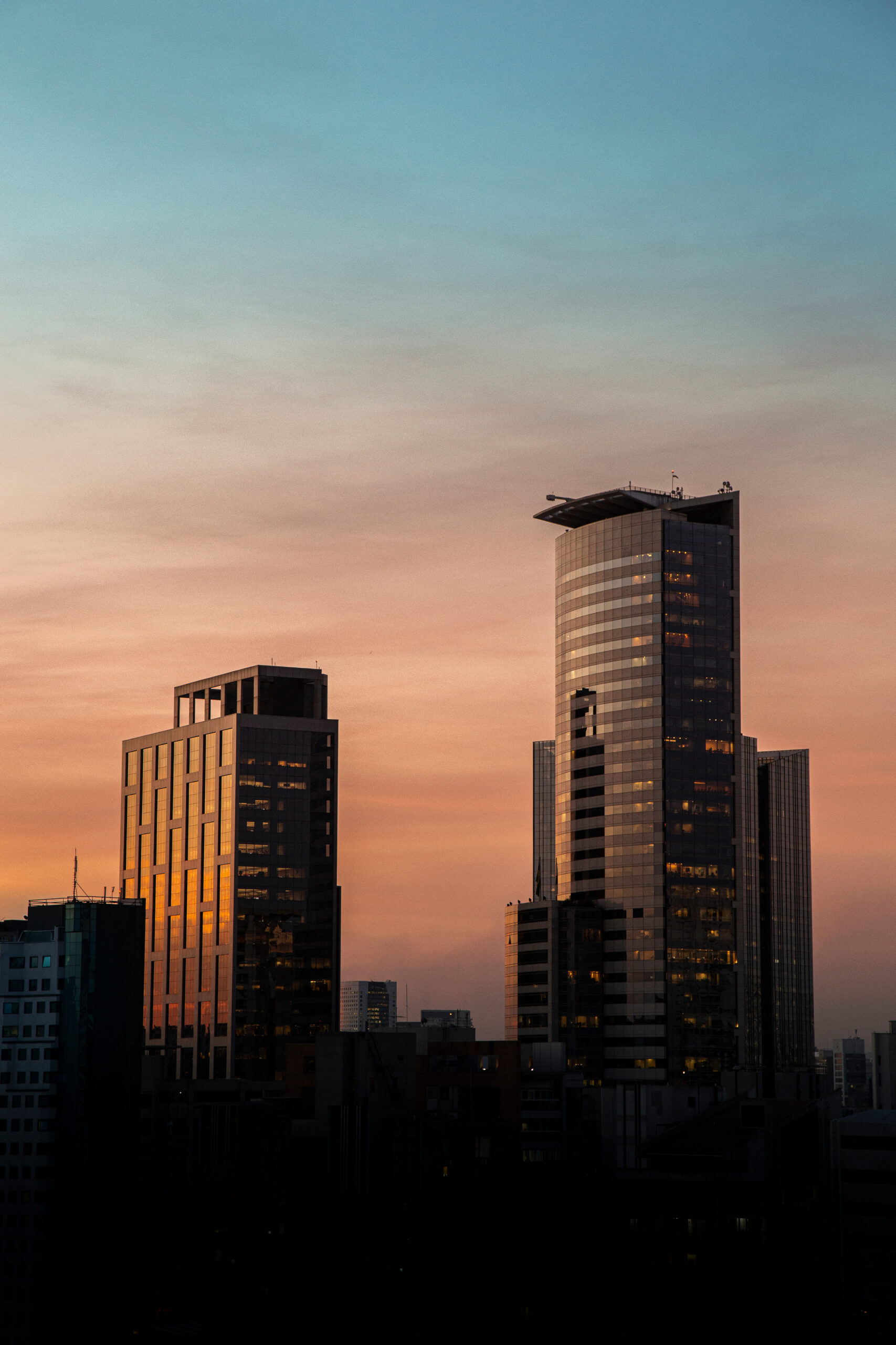 Prédios modernos de São Paulo ao pôr do sol, com céu em tons de azul e laranja, refletindo a luz dourada nas janelas espelhadas.